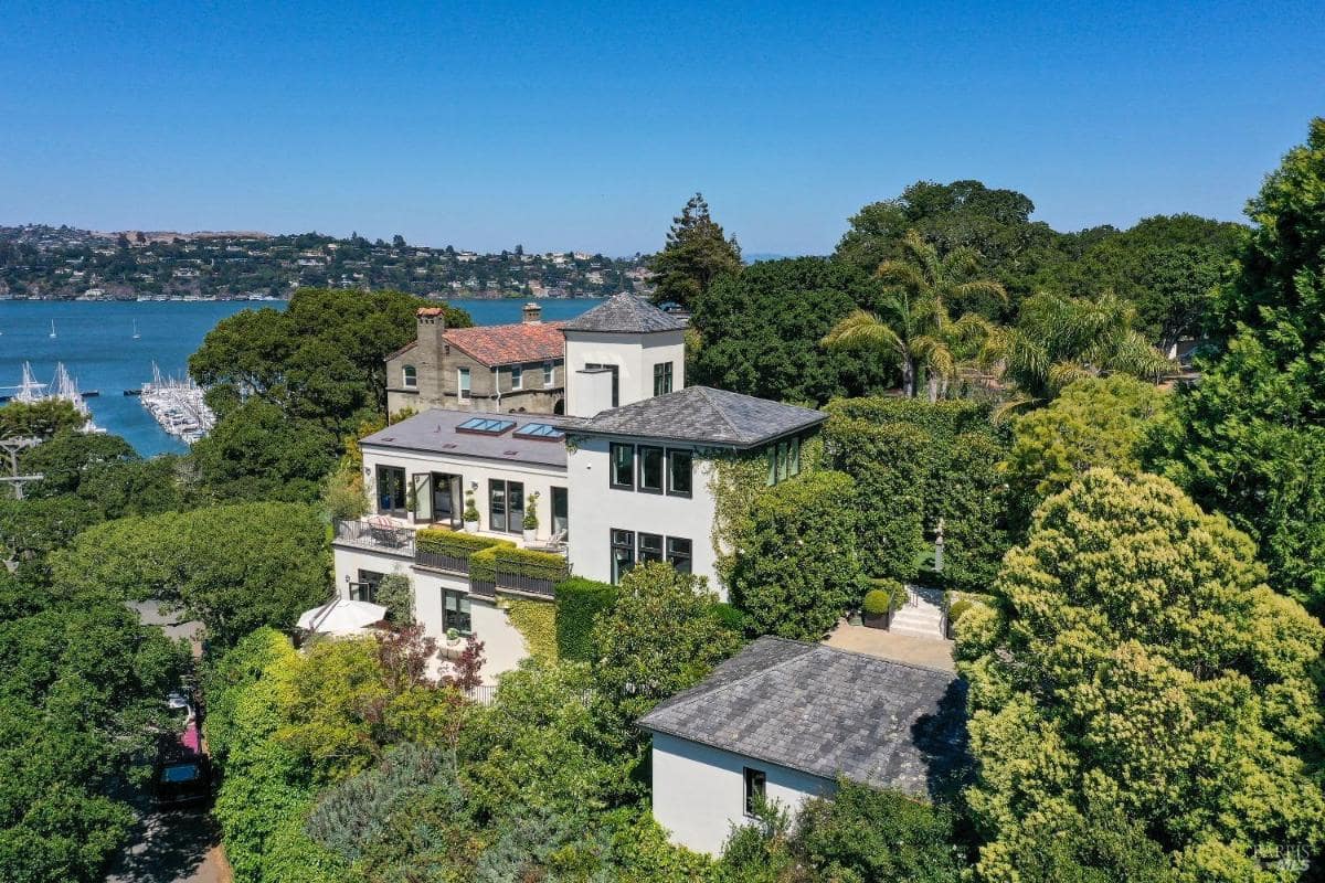 Aerial view of a multi-level house near a waterfront with surrounding greenery.