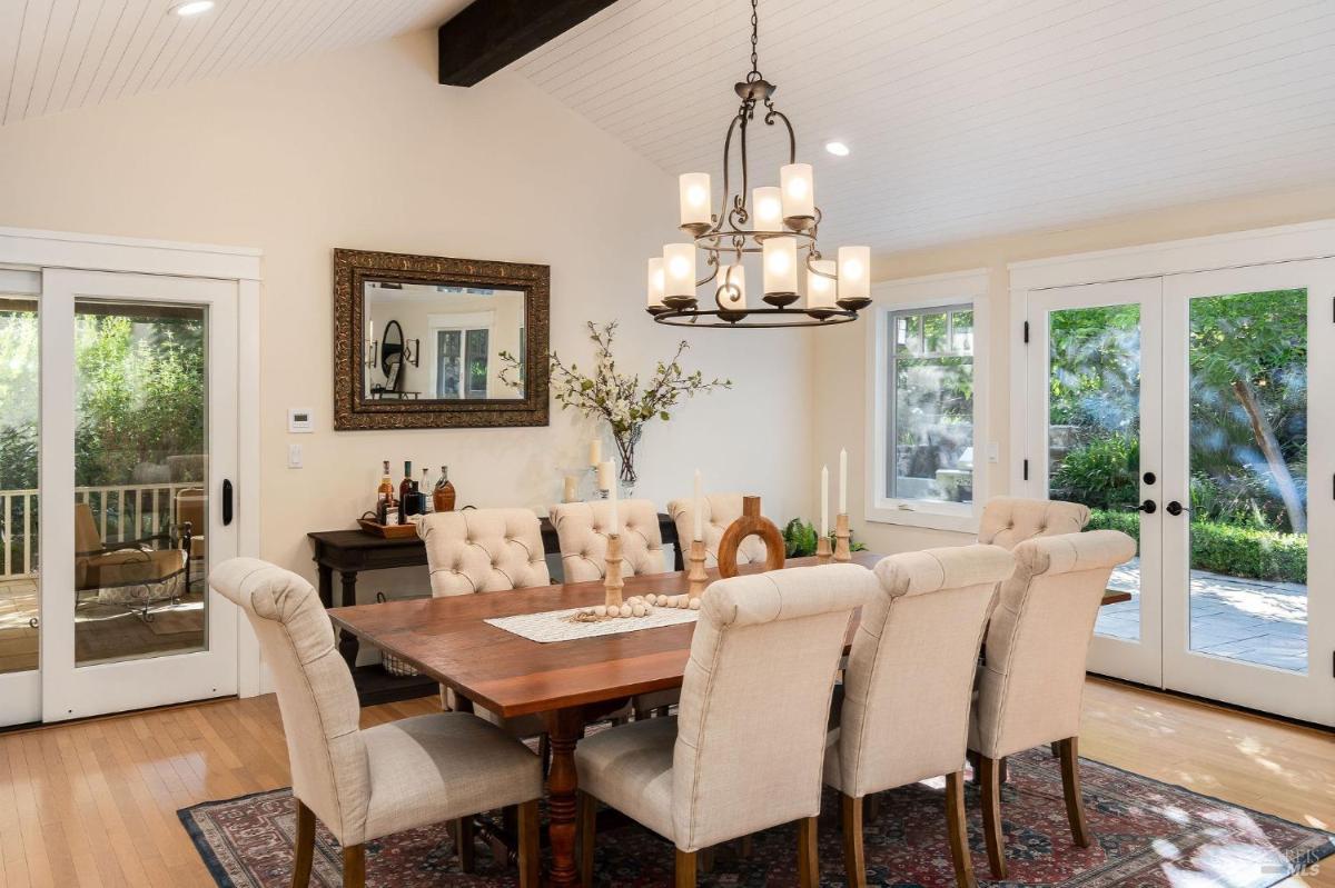 A dining room with a wooden table surrounded by chairs, featuring a chandelier and large windows.

