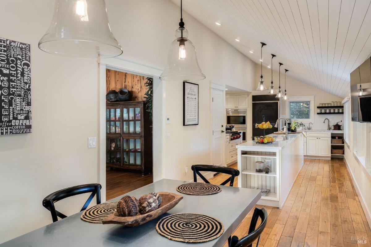 A dining area with a wooden table, black chairs, and a view of a kitchen with pendant lights.

