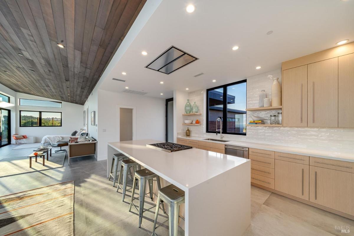 A combined kitchen and living space with large windows and a wood-paneled ceiling.