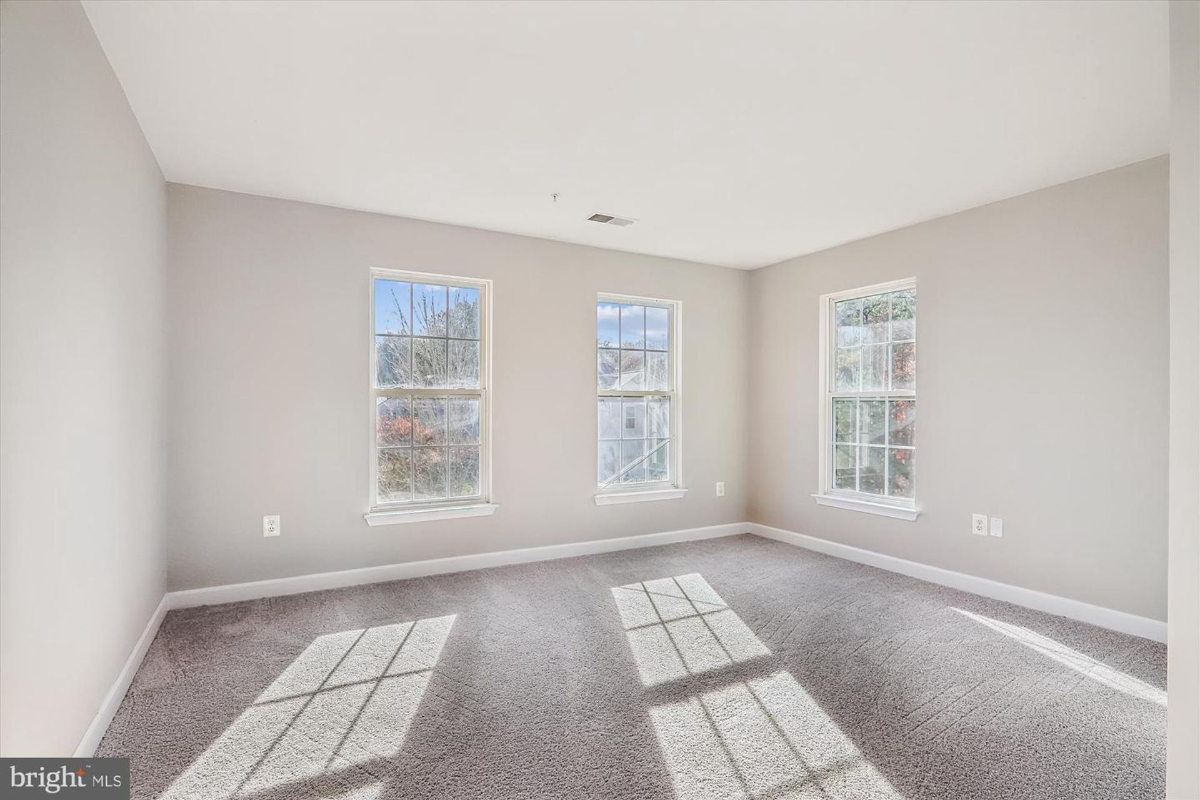 Empty bedroom with gray carpeting and three windows. The walls are painted a light neutral color.