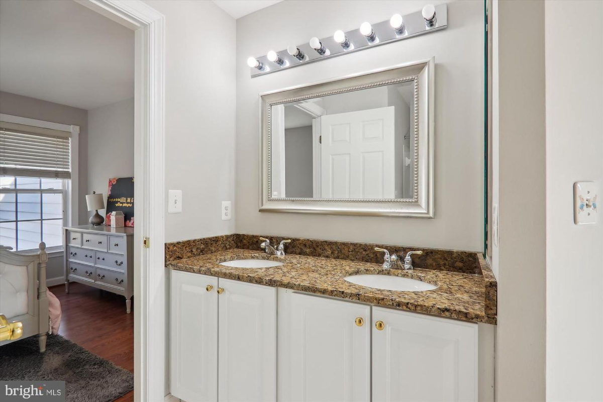 Bathroom with a double vanity, granite countertop, and a large mirror. A portion of a bedroom is visible in the background.