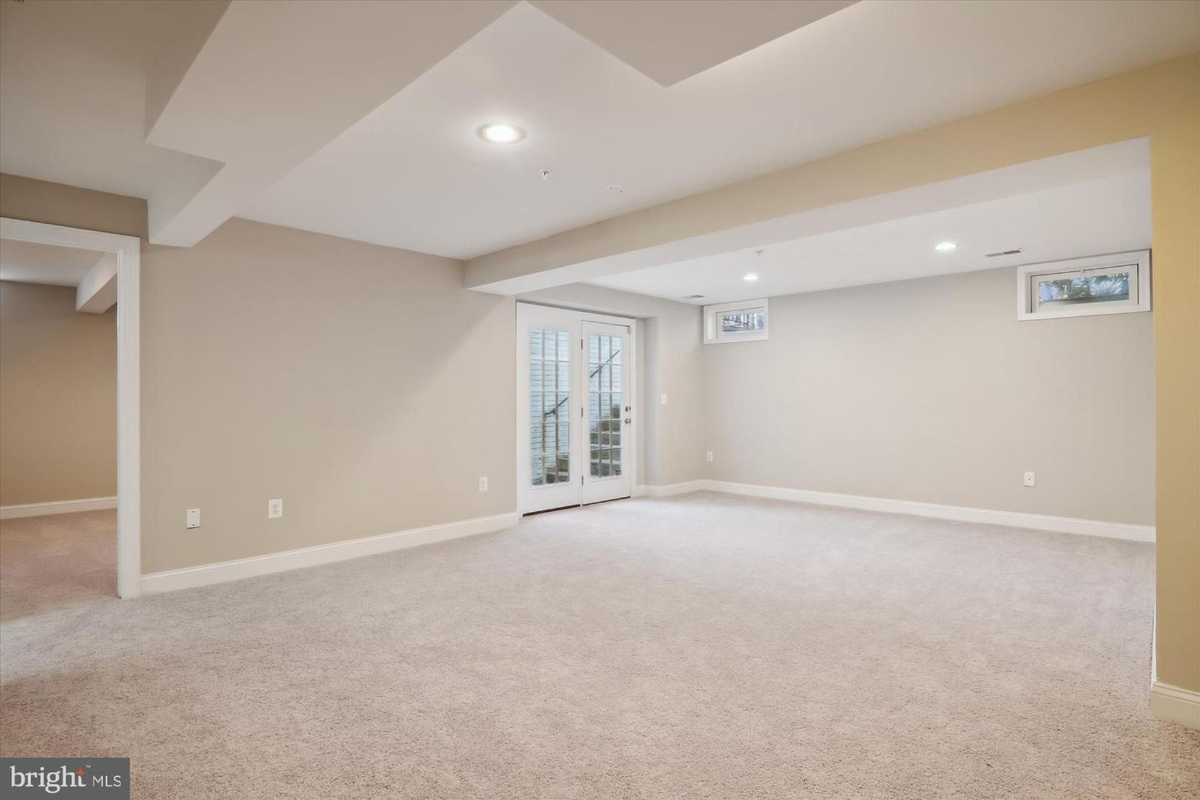 Large, empty basement room with gray carpeting and french doors leading to the outside. The walls are a light neutral color.