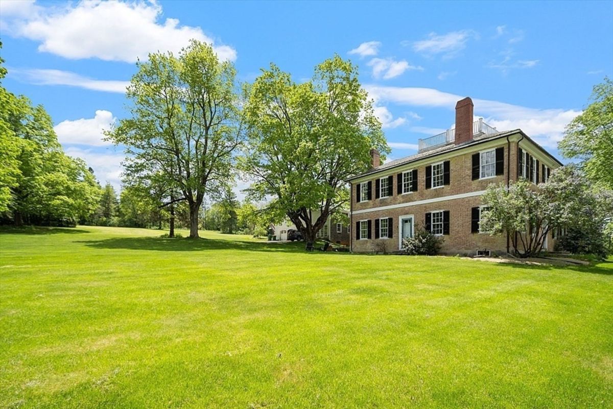 A large, tan brick colonial-style house with dark shutters and a rooftop balcony sits on a spacious green lawn