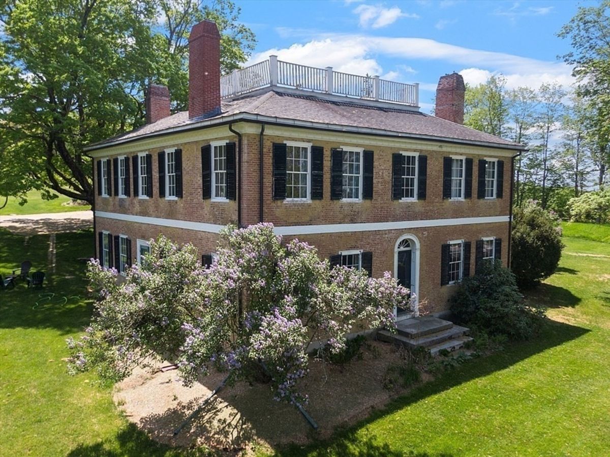 A large brick colonial house with a balcony sits on a grassy lawn.