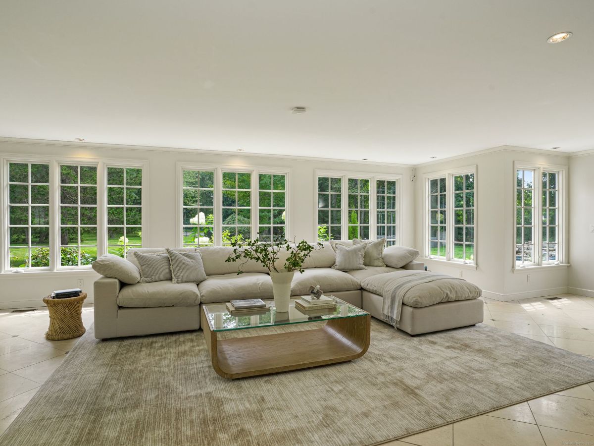 Sunlit lounge with wraparound windows, neutral tones, and a glass-top coffee table.