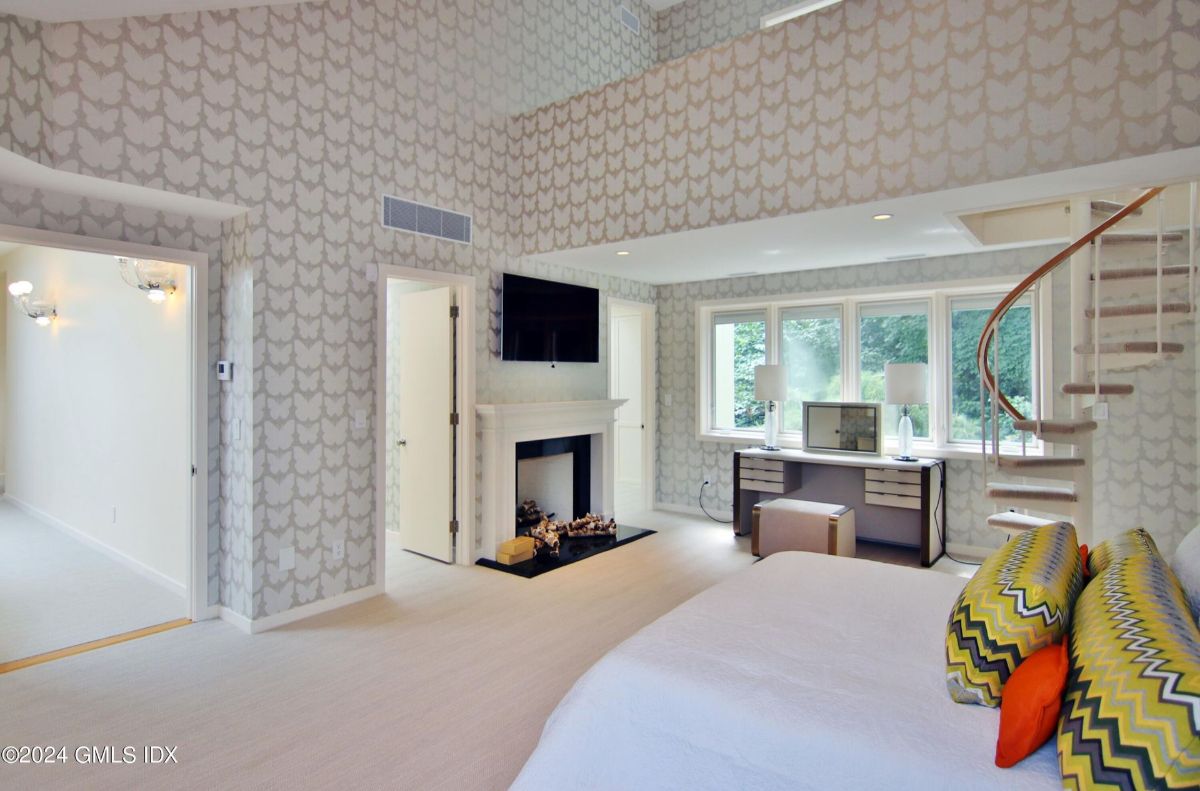 Bedroom features a modern fireplace, built-in TV, and an elegant workspace framed by large windows.