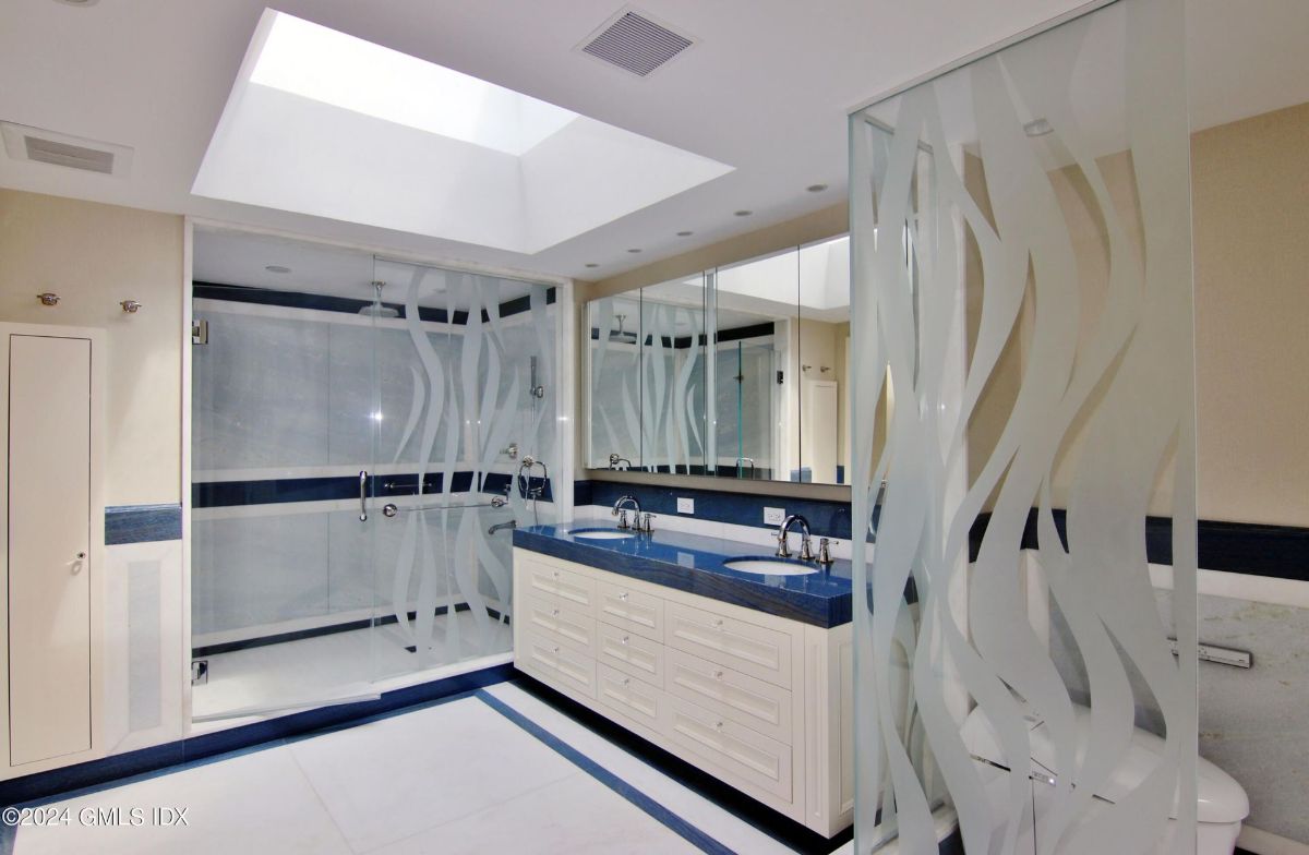 Bathroom featuring dual sinks with a bold blue countertop, frosted glass partitions, and a large skylight illuminating the modern shower area.