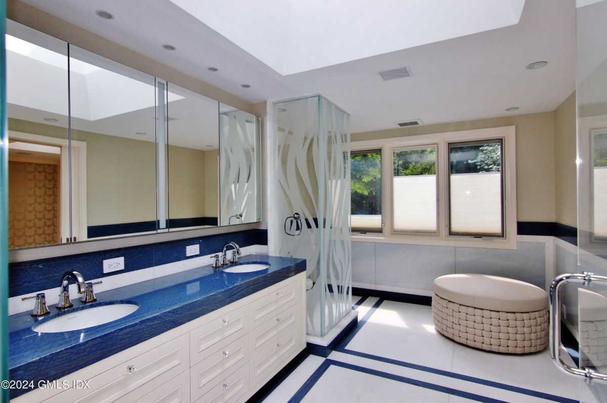 Bathroom showcases a vibrant blue countertop with dual sinks, frosted glass accents, and a stylish woven ottoman.