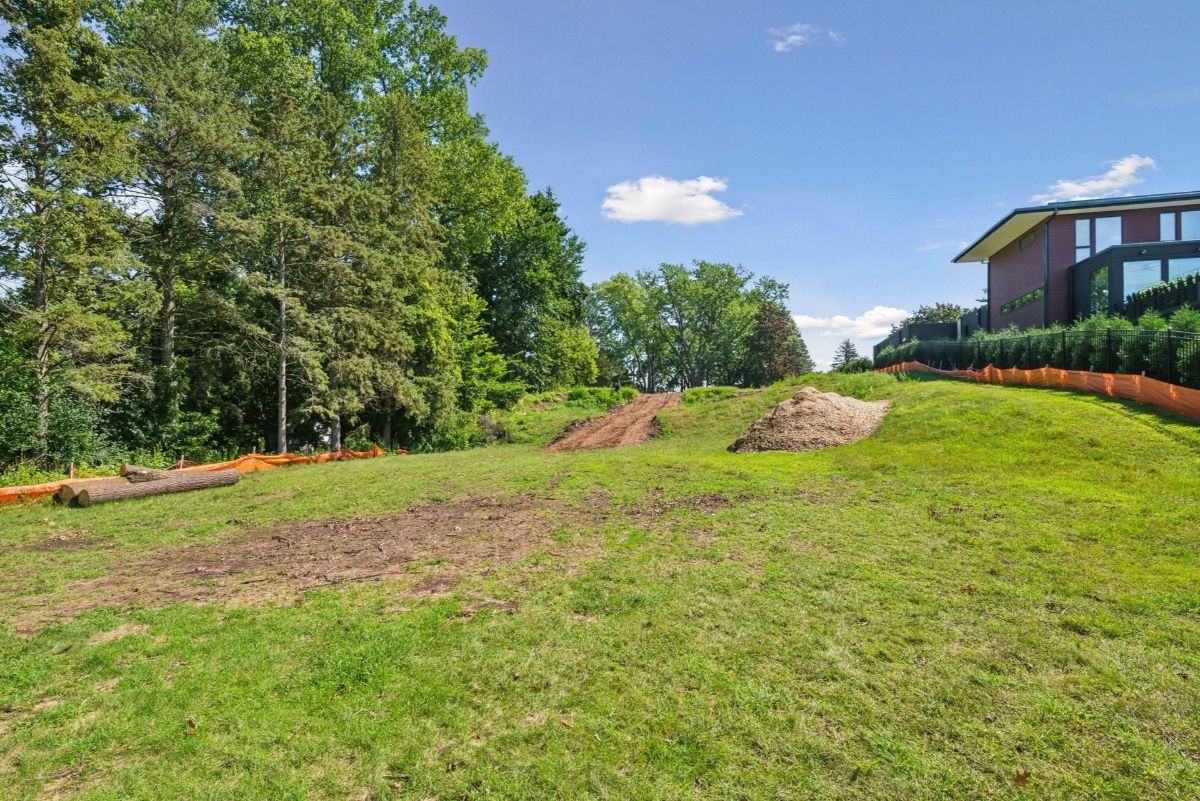 A sloped lawn with freshly cleared patches is bordered by tall trees and a modern home in the distance.