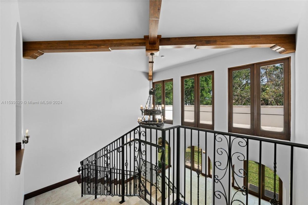 Second-floor landing with wood beam ceiling, iron chandelier, and large windows framing serene outdoor views.