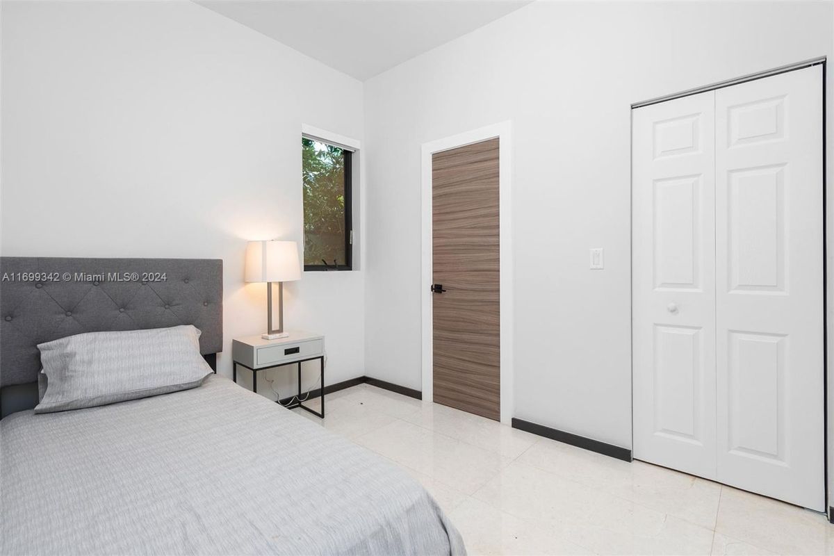 A minimalist bedroom layout includes a sleek gray upholstered bed, soft bedside lighting, and a window inviting natural greenery.