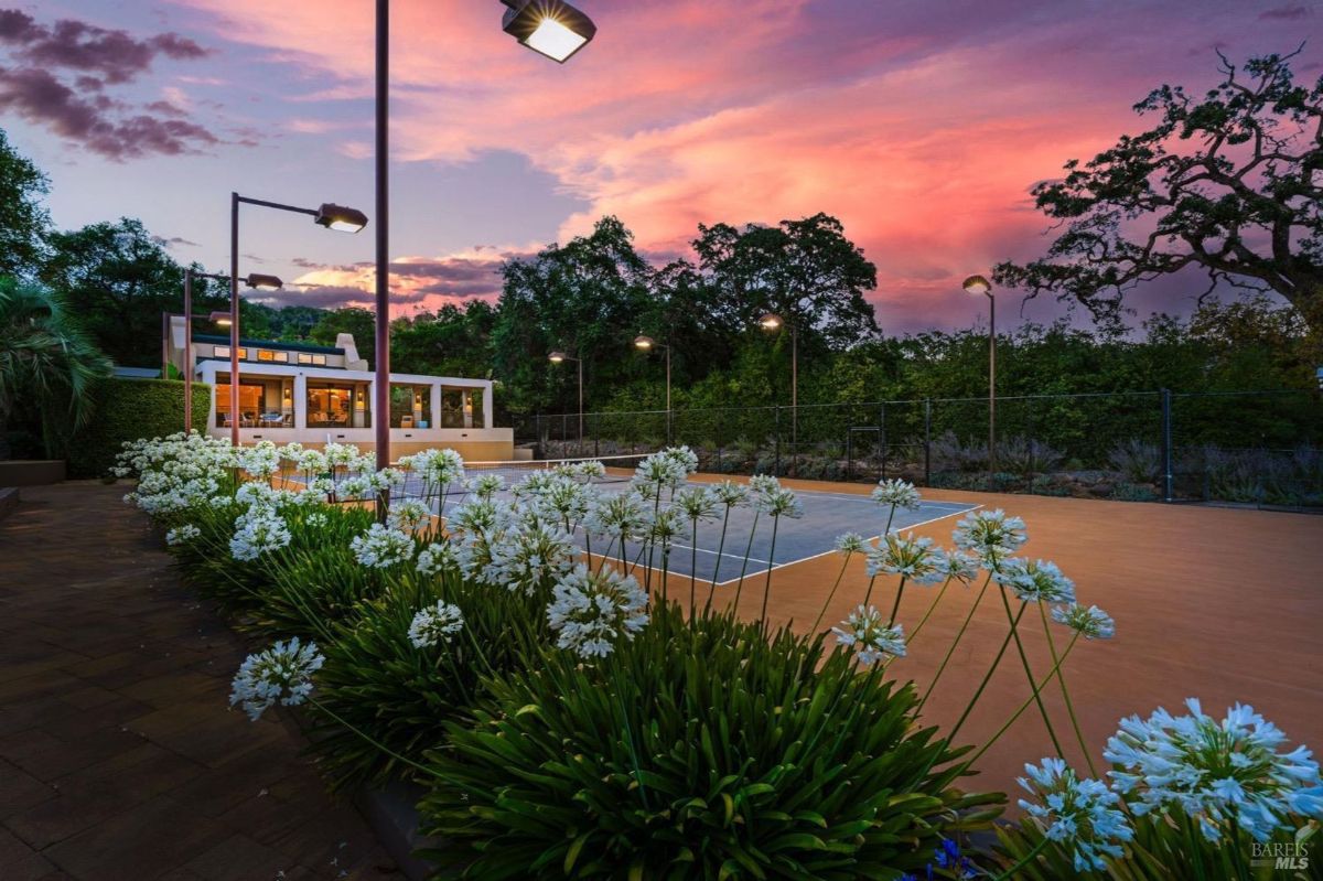 Lit pathways and blooming flowers frame the tennis court, set against a vibrant evening sky.