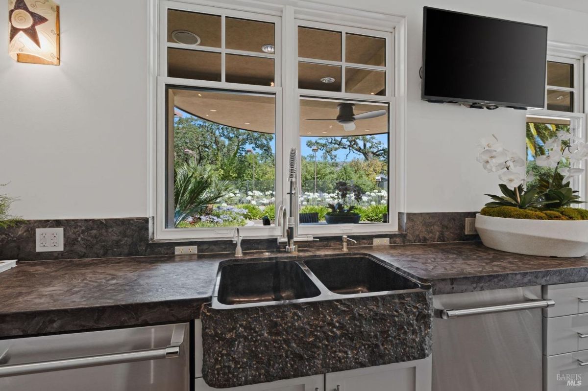 Farmhouse-style sink framed by rich countertops and picturesque garden views through large windows.