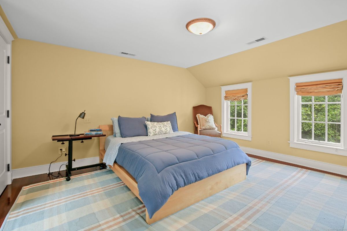 Sunlit bedroom with cozy blue bedding, warm yellow walls, and windows offering serene views of greenery.