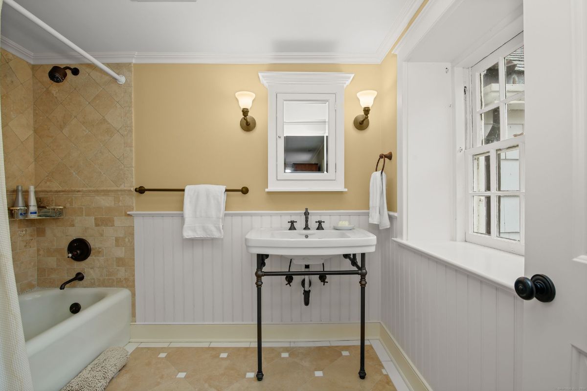 Bright bathroom with a pedestal sink, wainscoting, and a tub with natural stone tile accents.