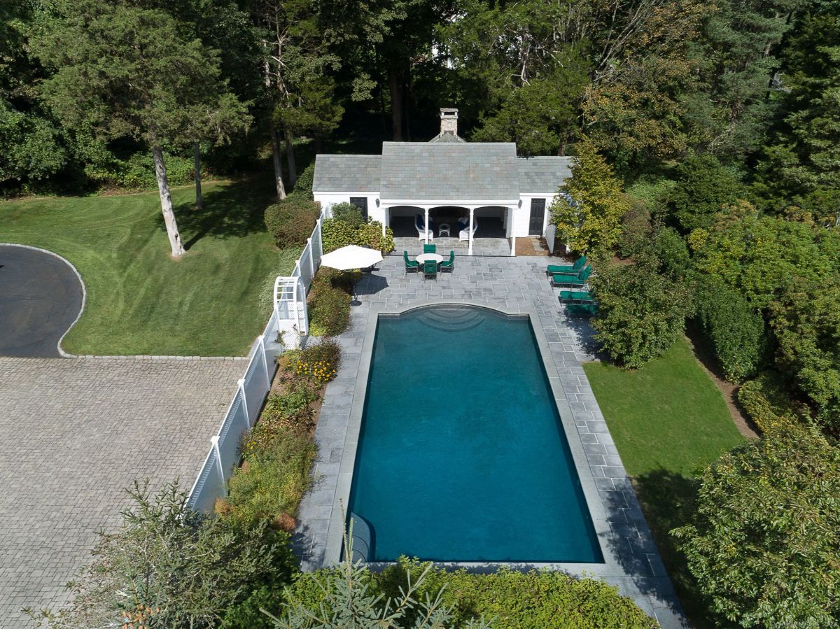 Elegant pool area with a slate patio, pool house, and lush surrounding greenery.