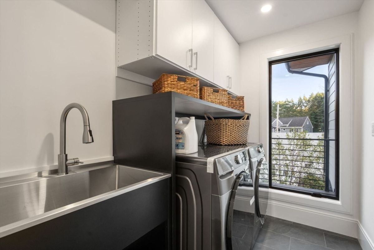 Laundry room equipped with sleek cabinetry, a stainless steel utility sink, and modern washer and dryer units, complemented by a window view.