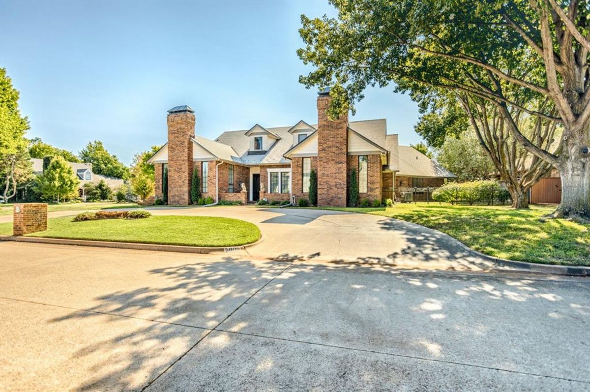 Brick-clad estate featuring dual chimneys and a circular driveway, surrounded by lush greenery.