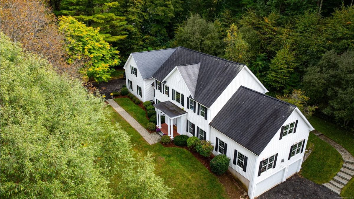 An aerial view of a large white two-story house with a two-car garage, surrounded by lush green trees.