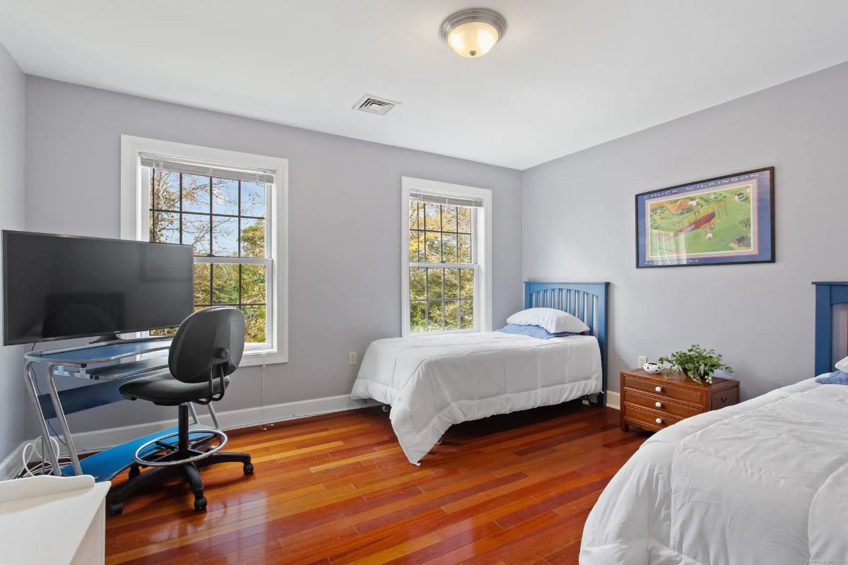 A bedroom with hardwood floors, light gray walls, and two twin beds, featuring a desk with a computer and windows offering views of trees.