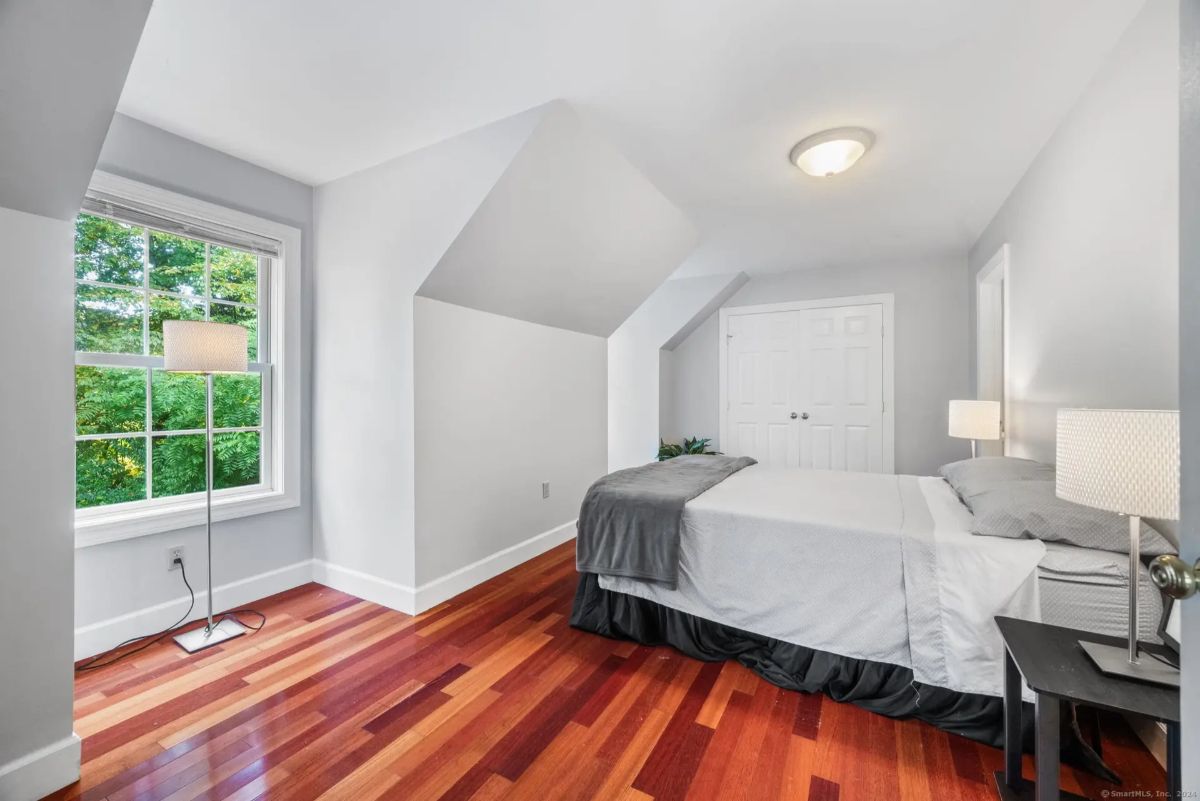 Simply decorated bedroom with hardwood floors, gray walls, and a double bed, featuring a large window offering a view of trees.