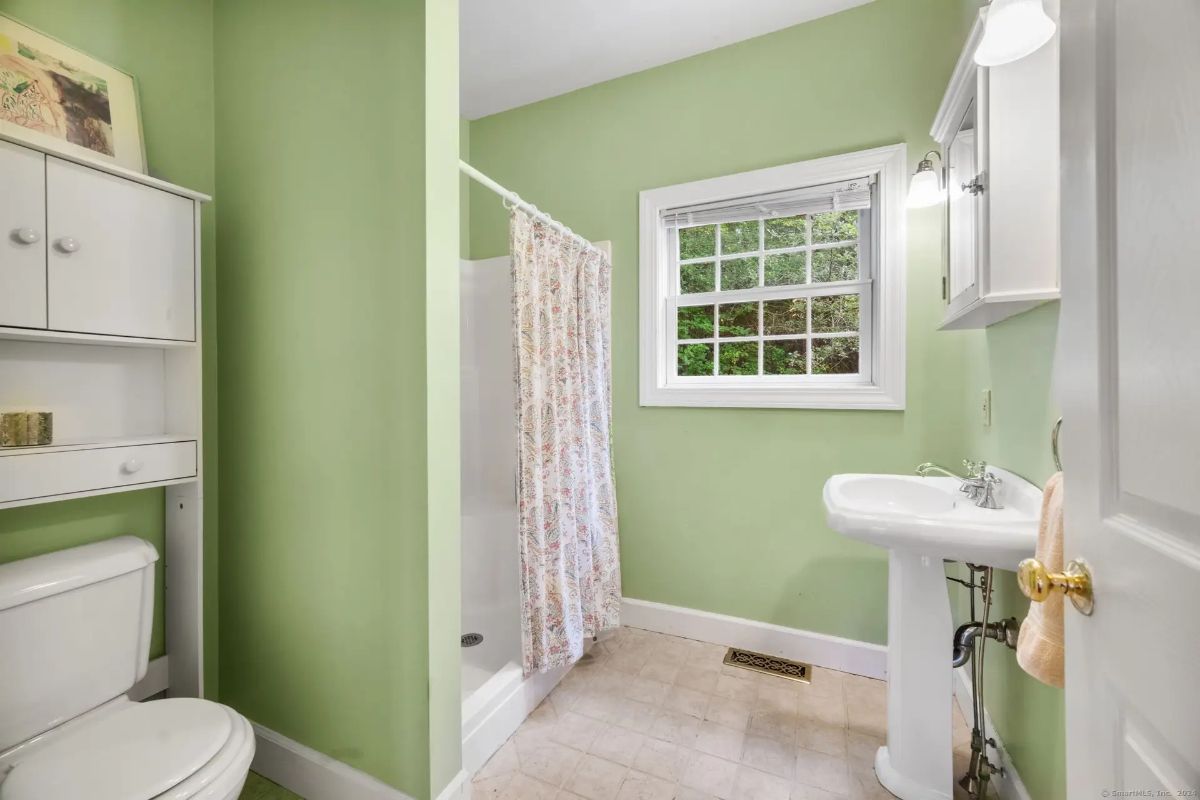 Light green bathroom with a pedestal sink, a shower stall with a floral curtain, and a window overlooking trees.