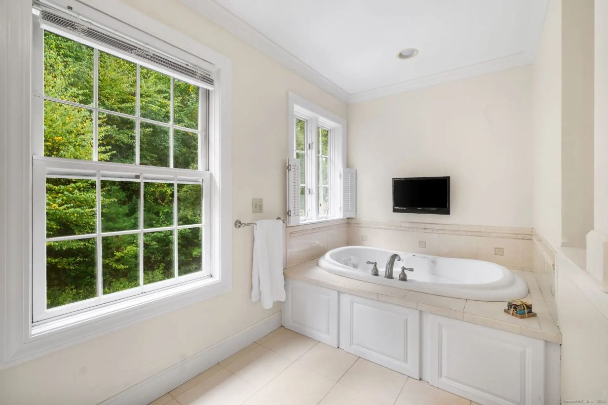 A luxurious bathroom with a large soaking tub, a flat-screen TV, and windows overlooking lush greenery.