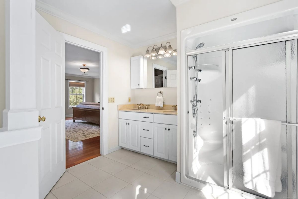 A bright and spacious bathroom with a double vanity, a large walk-in shower, and an open doorway leading to a bedroom. 