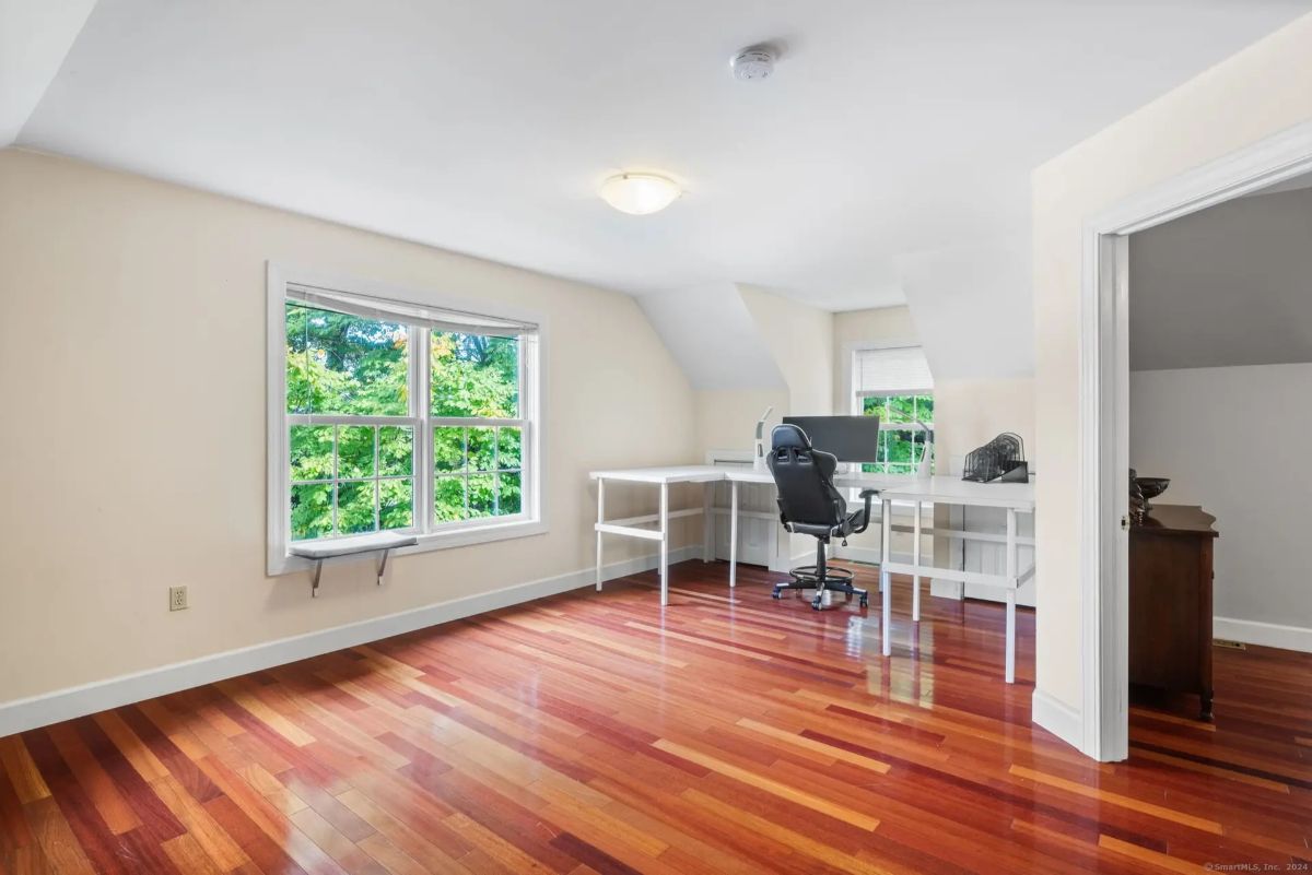 An office or study with hardwood floors, a desk and chair, and a large window overlooking trees.