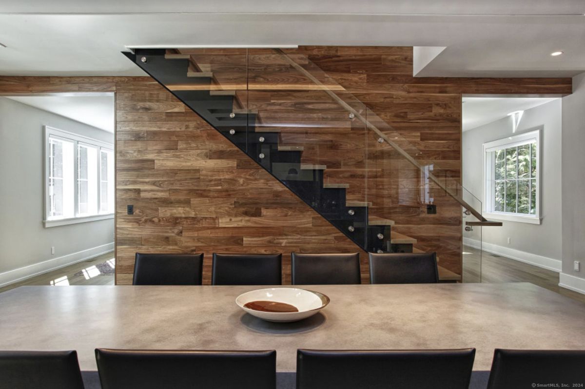 Dining area sits adjacent to a modern staircase with glass railing and a rich wooden accent wall for a warm and contemporary touch.