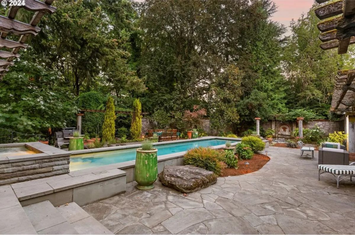 A long rectangular pool surrounded by a stone patio and lush greenery.