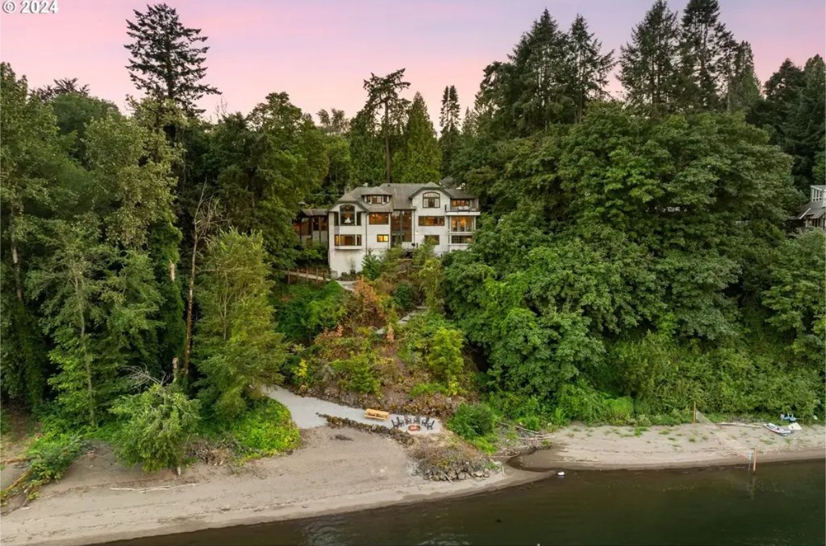 A multi-level white home nestled on a forested hillside along a riverbank.