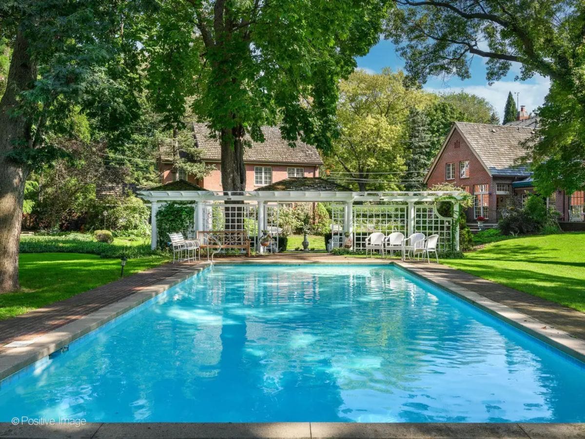 Large, rectangular swimming pool is surrounded by greenery and shaded by mature trees.