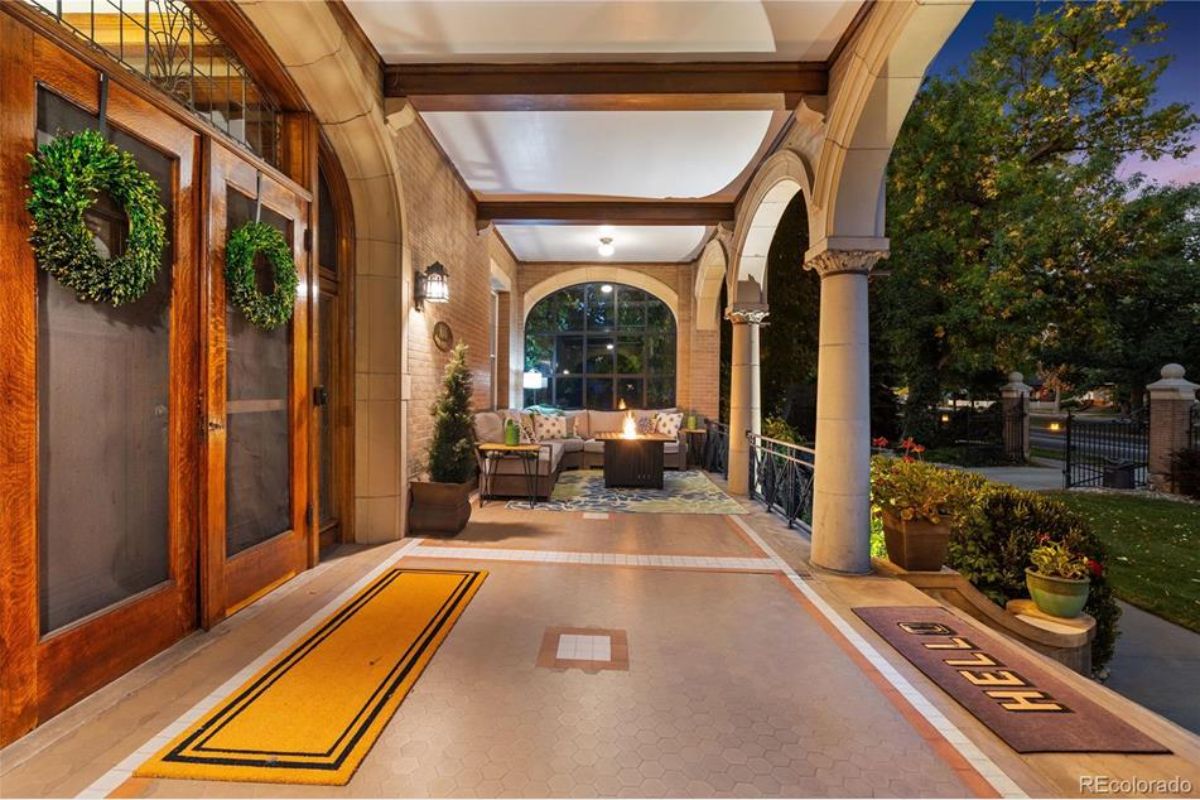 The illuminated porch of the Sharp Mansion showcases its timeless charm with arched columns, intricate tile flooring, and a warm, inviting seating area centered around a fire feature. 