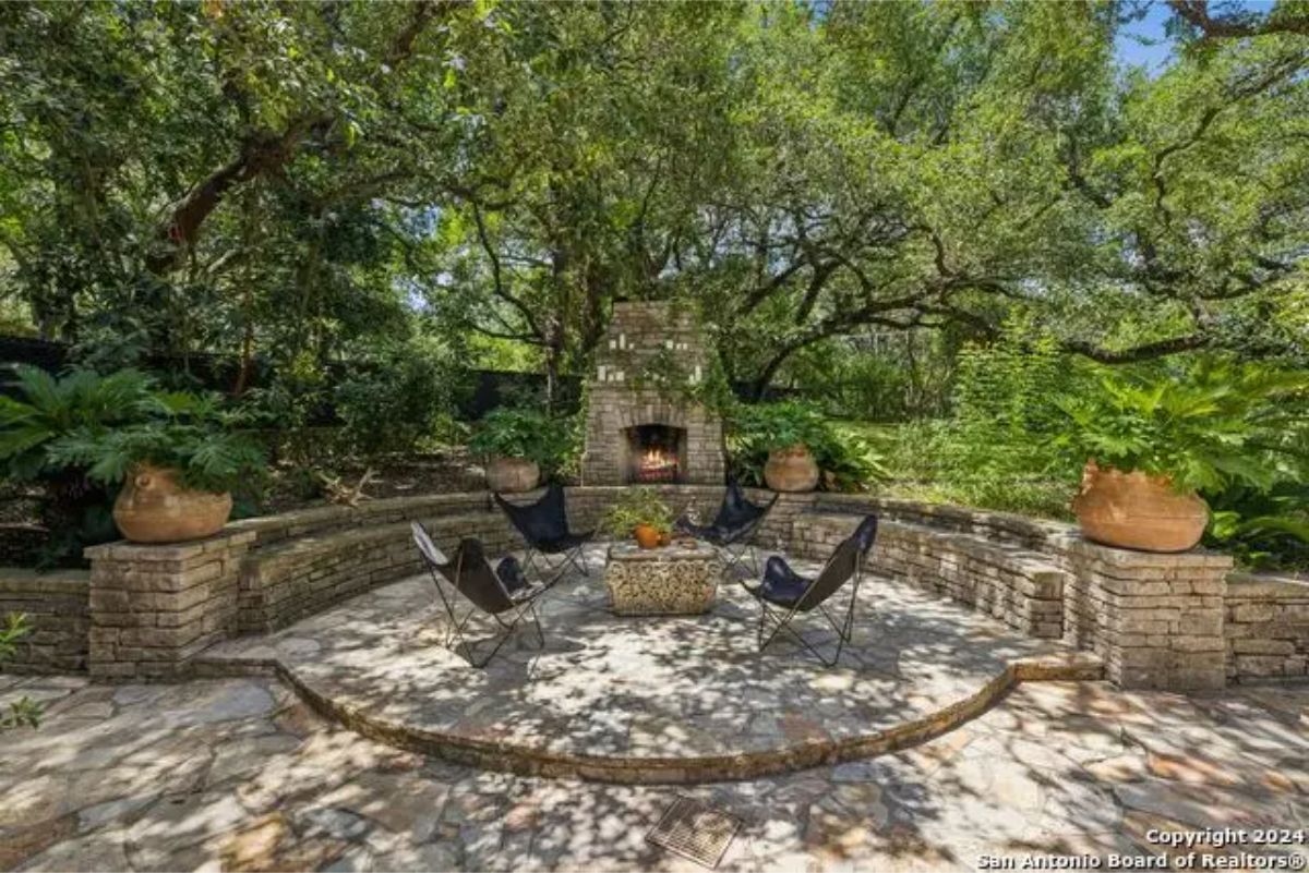 A stone fireplace stands as the centerpiece in a circular outdoor seating area surrounded by lush greenery.