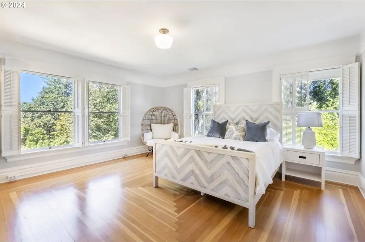 Bedroom with light walls and natural wood floors features large windows offering views of lush green trees.