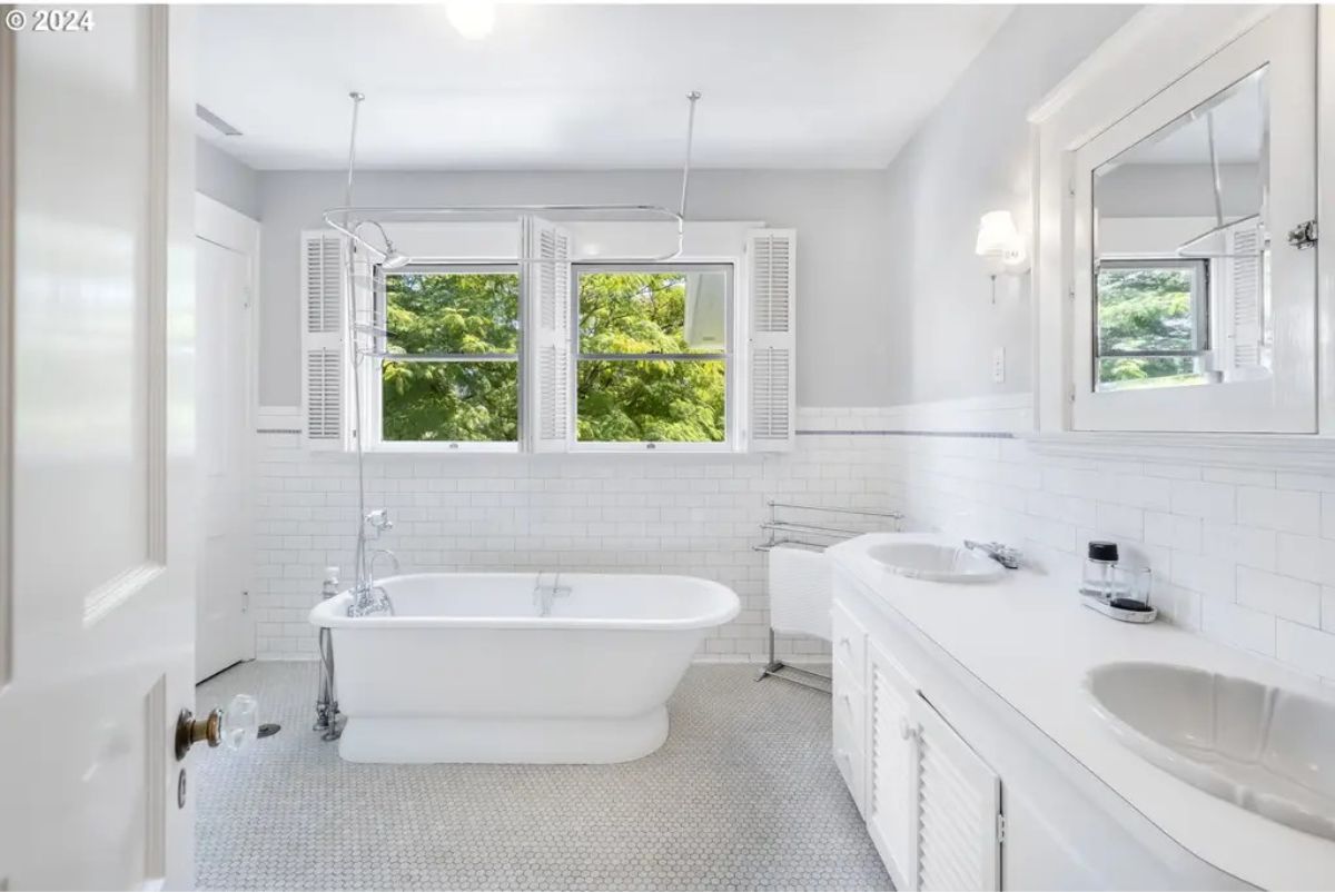 A freestanding clawfoot tub positioned near two large windows with shutters, providing views of greenery.