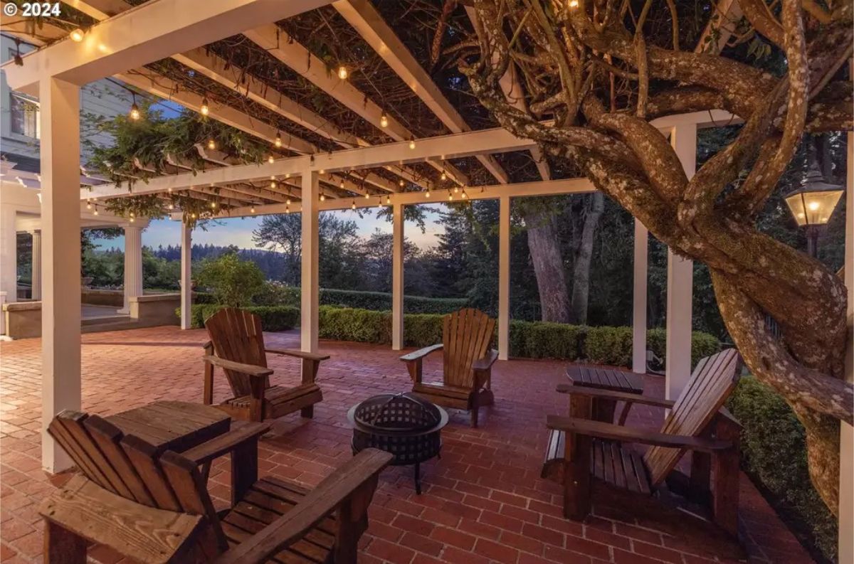 Wooden chairs are arranged around a firepit on a red brick patio beneath a pergola draped with greenery.