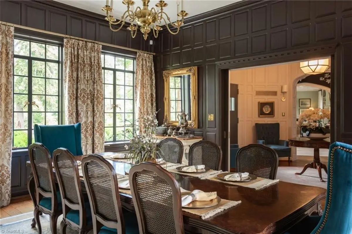 Dining room is defined by dark wood paneled walls, contrasted by large windows with patterned drapes.