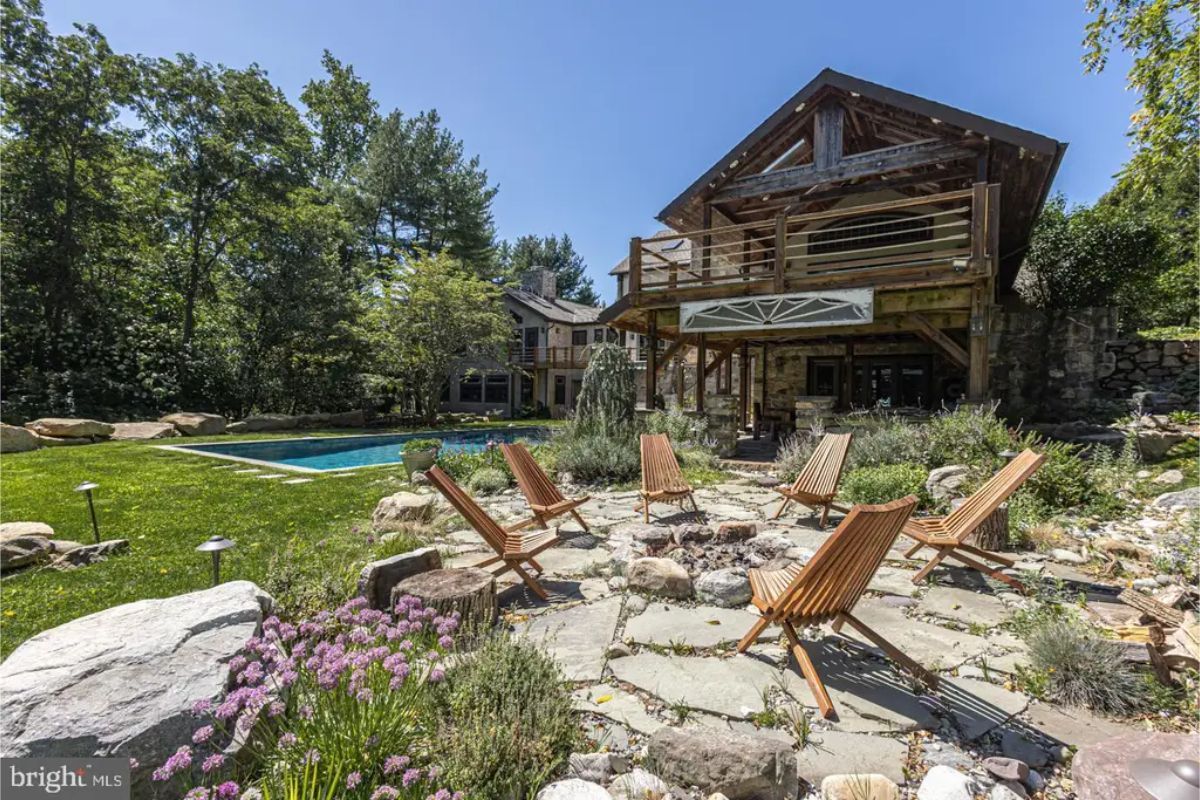 Wooden chairs encircle a fire pit on a stone-paved patio surrounded by landscaping.