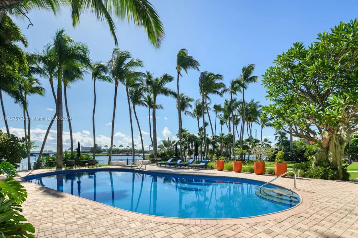 Curved swimming pool surrounded by a paved deck and tropical landscaping, including palm trees and vibrant greenery.