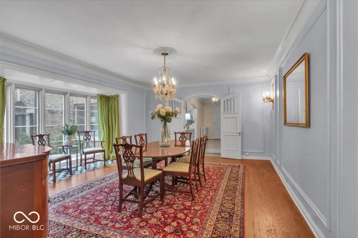 The dining room showcases a long wooden table surrounded by carved chairs on a vibrant red patterned rug. 