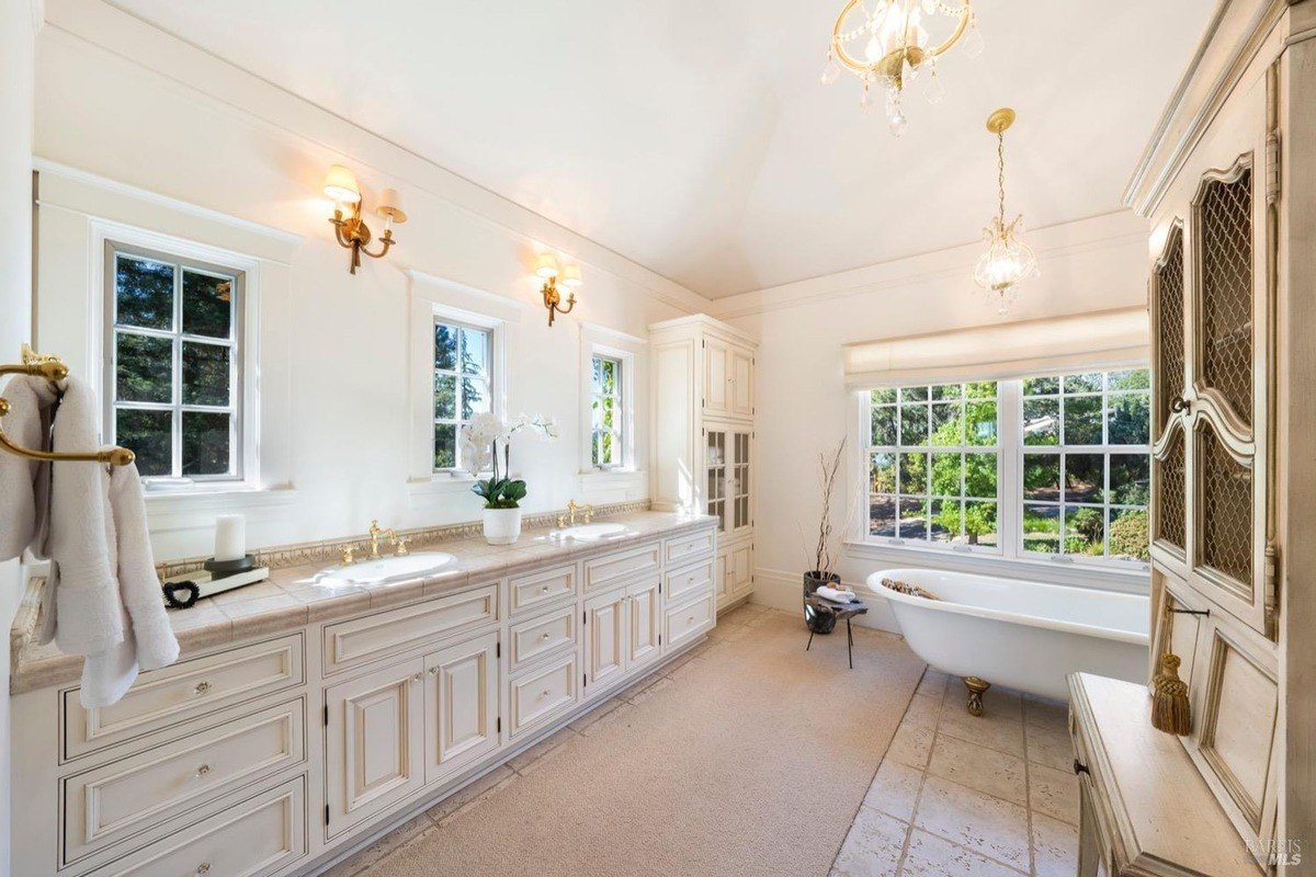 Bathroom has white cabinets, a clawfoot tub, and large windows overlooking trees.