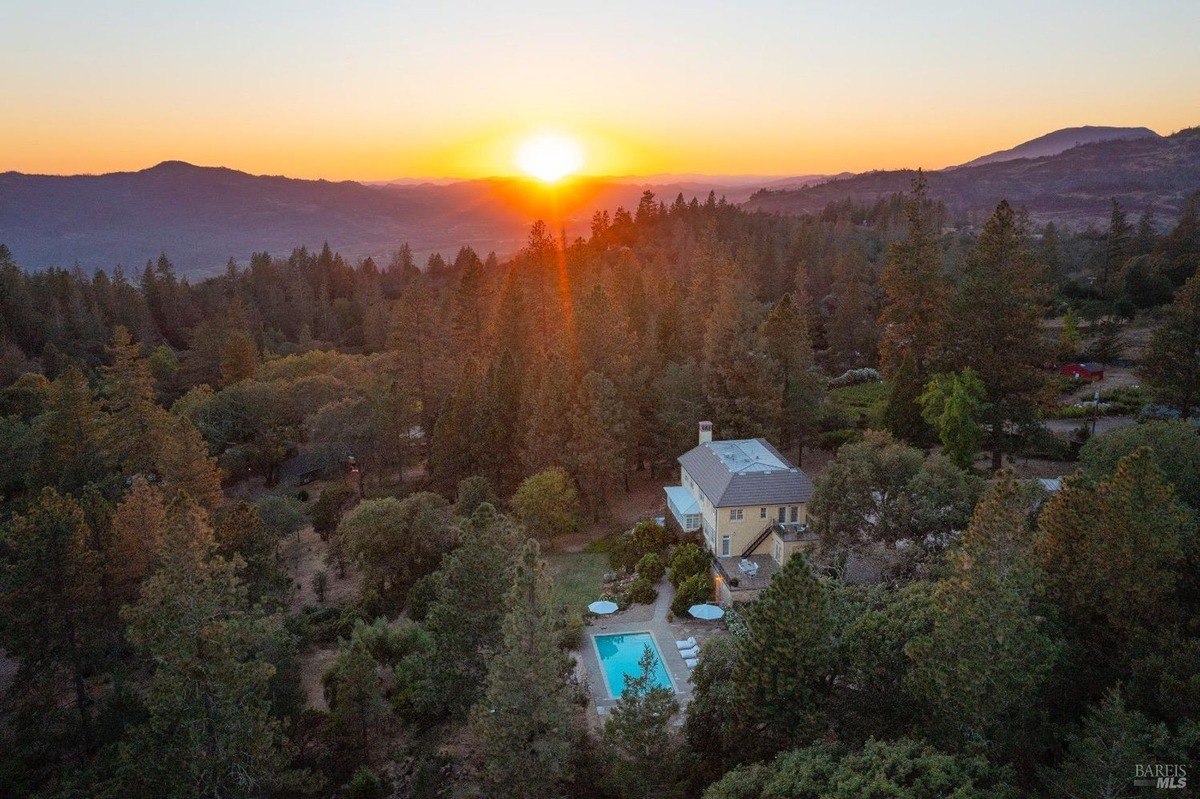 House is nestled in a wooded area with a pool, overlooking a valley at sunset.