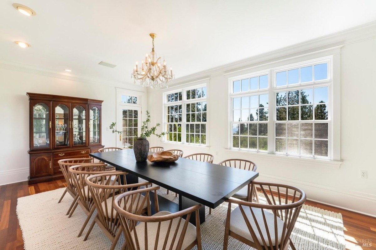 Dining room has hardwood floors, a large black table with wooden chairs, and a view of trees.