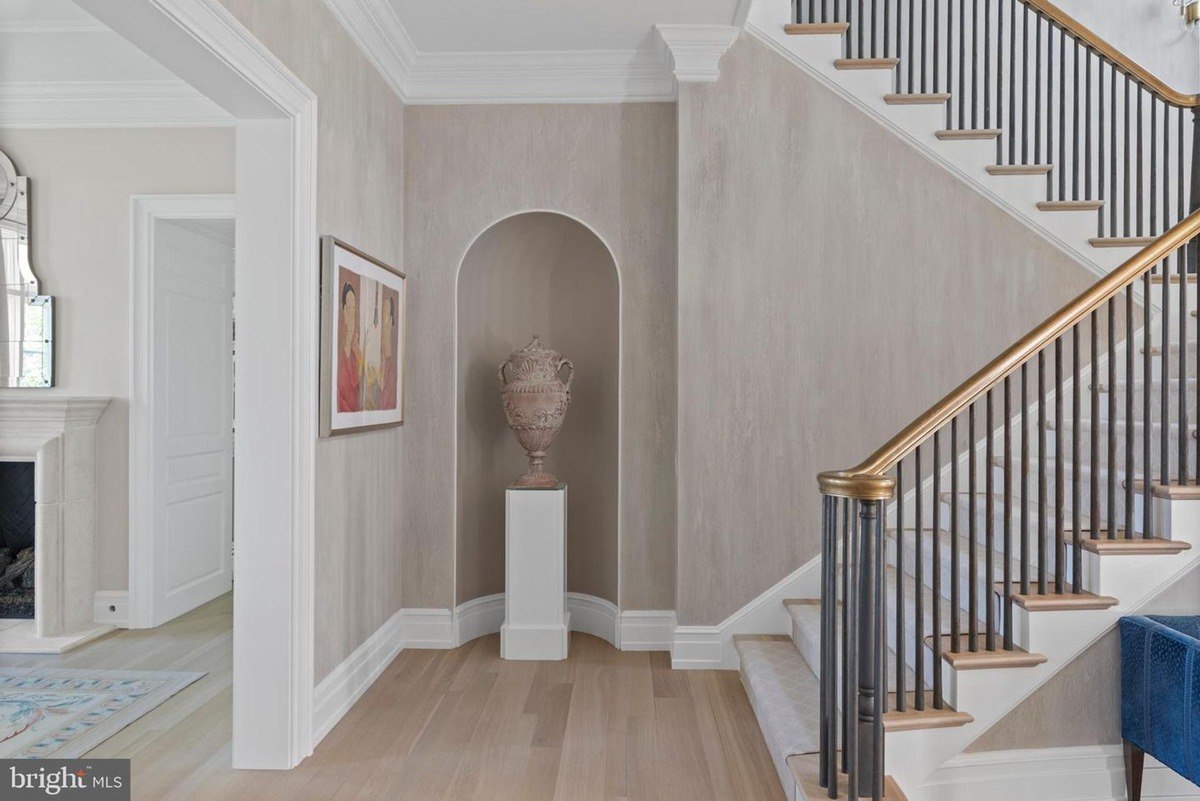 Elegant entryway features a staircase with a gold railing, neutral-toned walls, and a large urn on a pedestal.