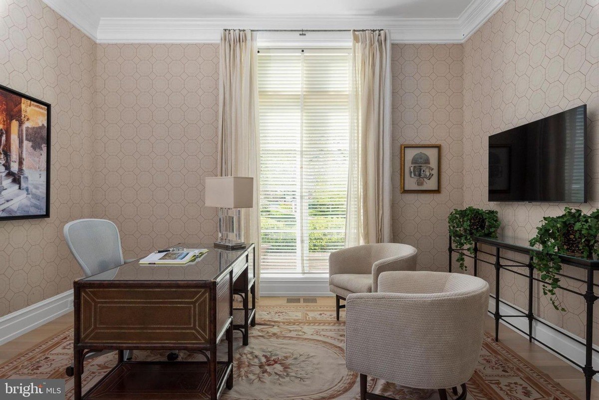 Home office features neutral-toned walls, a wood desk, comfortable armchairs, and a large window.