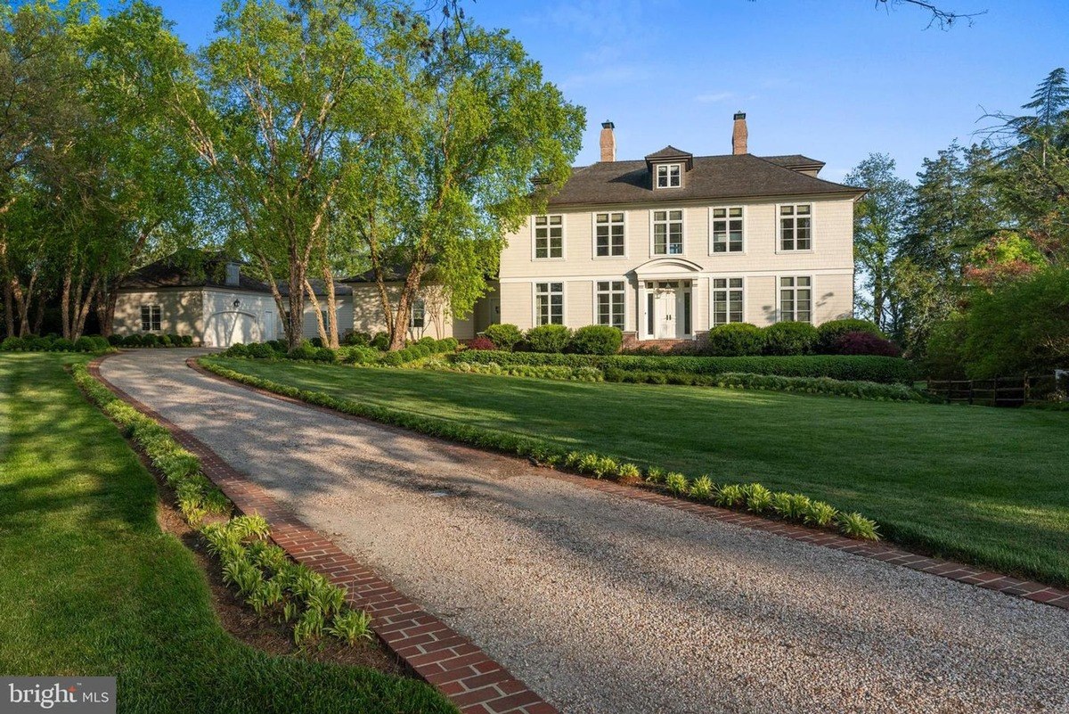 Long gravel driveway curves toward a large, light-colored, two-story house surrounded by green lawns and trees.