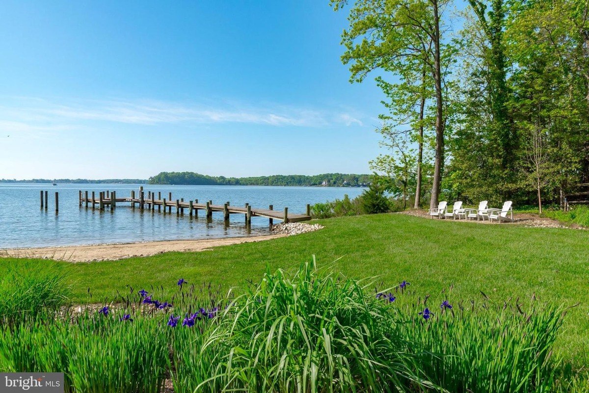 Landscaped lawn overlooks a calm body of water with a dock, featuring several white Adirondack chairs.